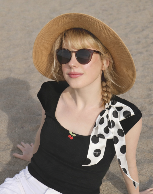 Woman sitting on sand wearing a straw hat, black top, white pants, and sunglasses.
