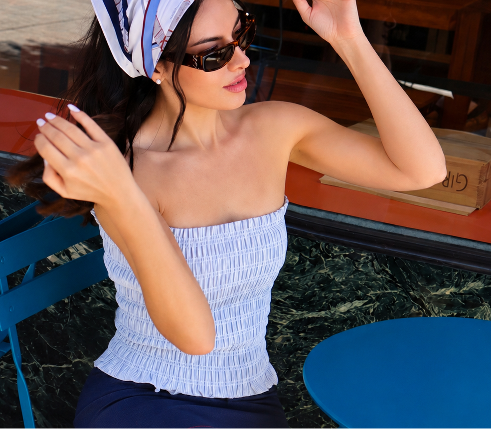 A woman sitting on a blue folding chair at an outdoor cafe, wearing a light blue and white vertically striped smocked tube top, a navy blue skirt, a blue and white patterned headscarf, and black sunglasses.