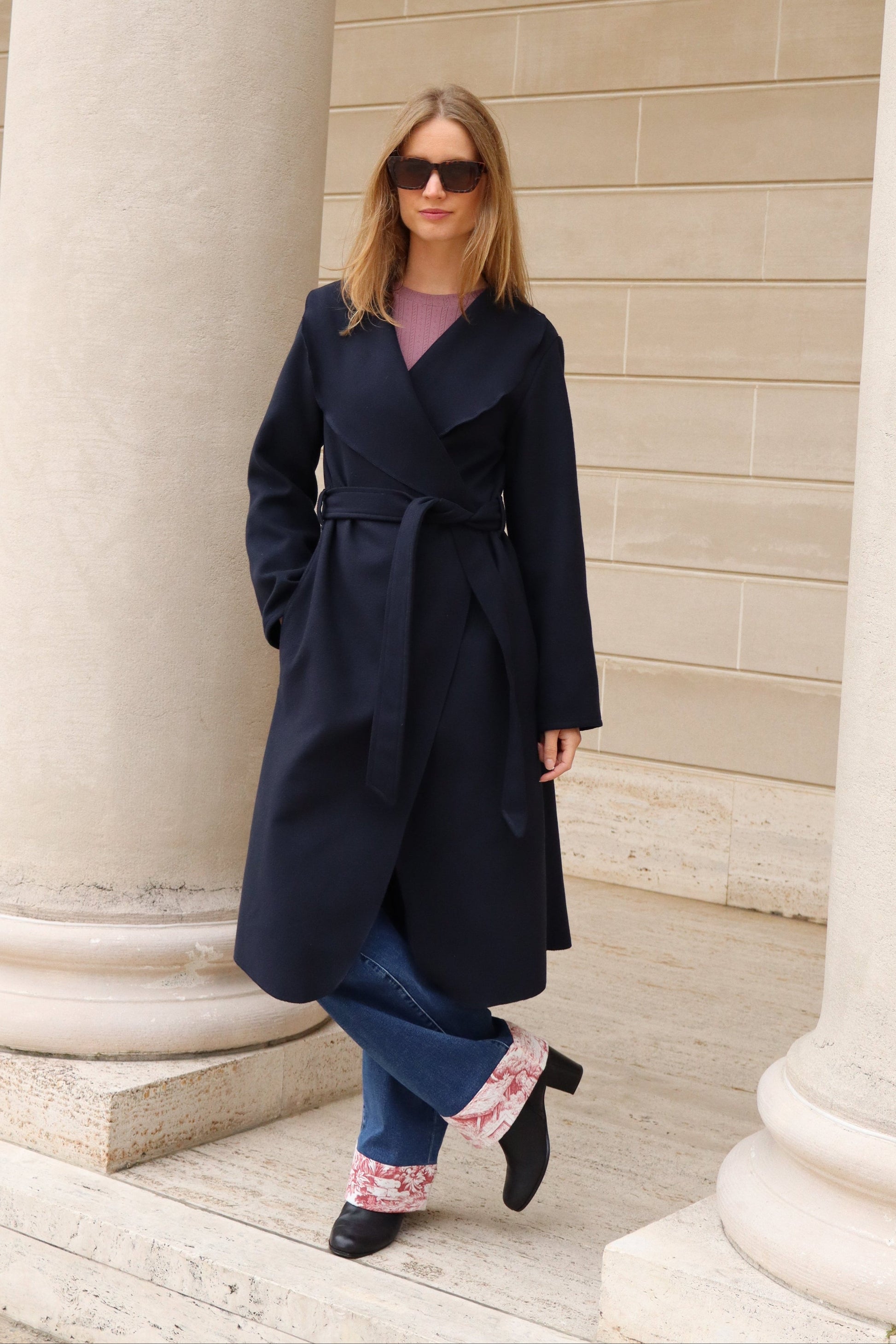 Woman wearing a navy coat and blue and red jeans with black boots against marble column