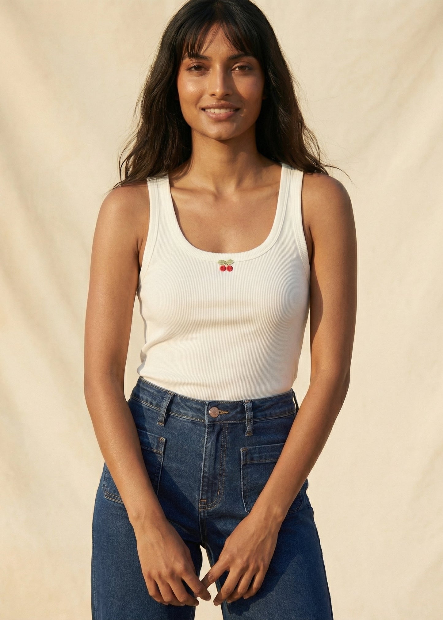 Woman wearing a white tank top with cherry design and blue jeans against a beige background