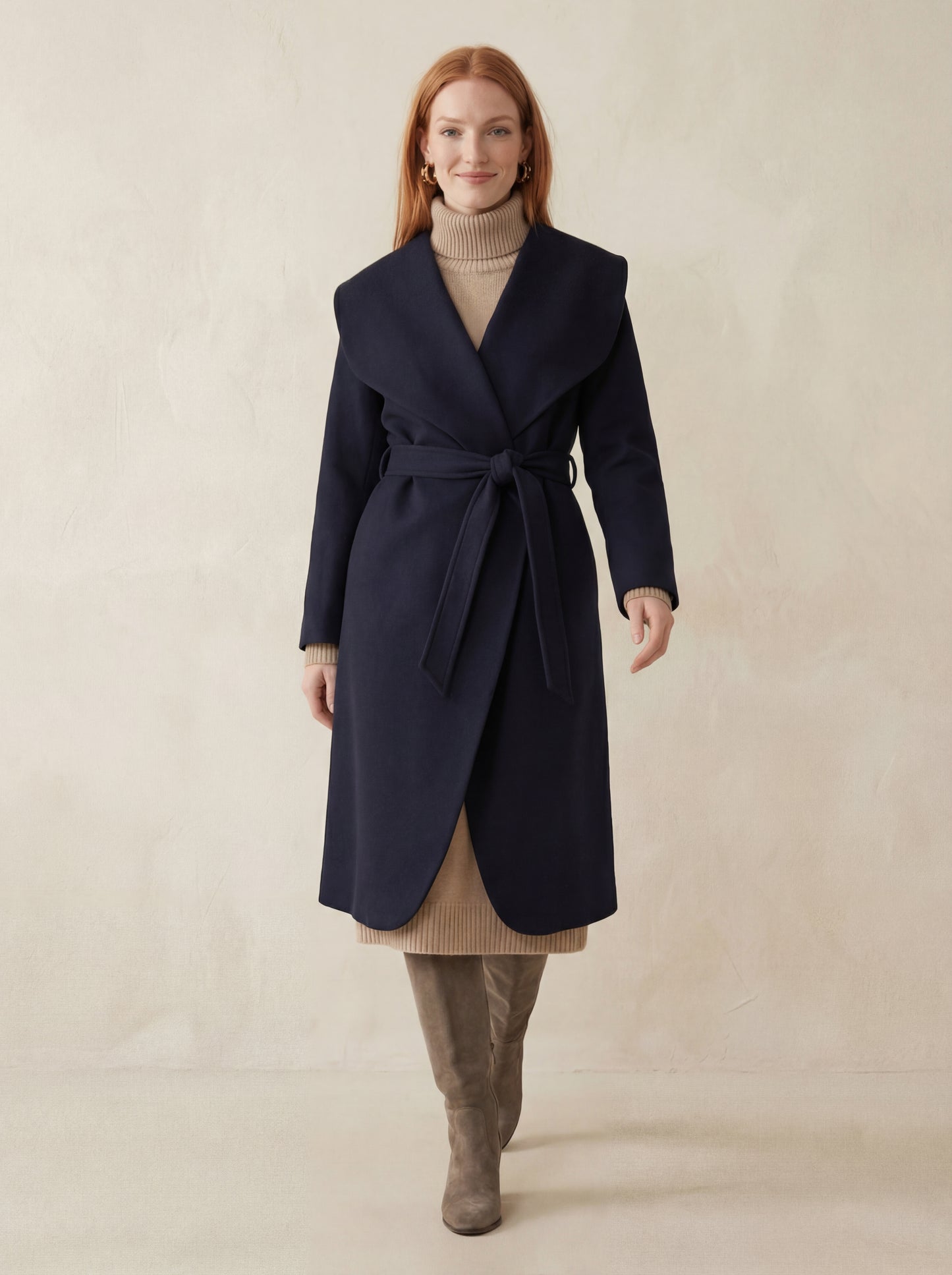 Woman wearing a navy coat with a beige sweater and brown boots on a plain background