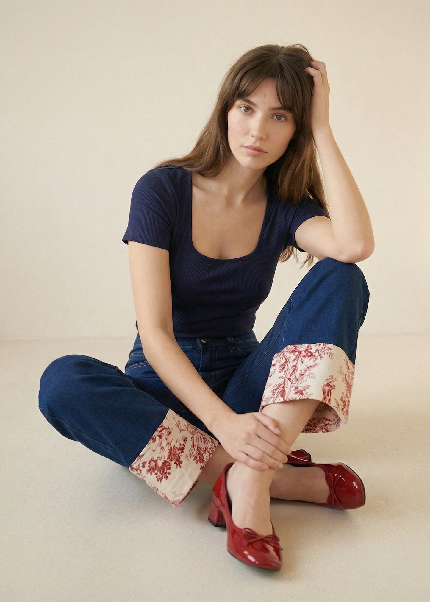 Woman sitting on the floor wearing a navy blue top and wide-leg pants with floral cuffs, red shoes, against a beige background.