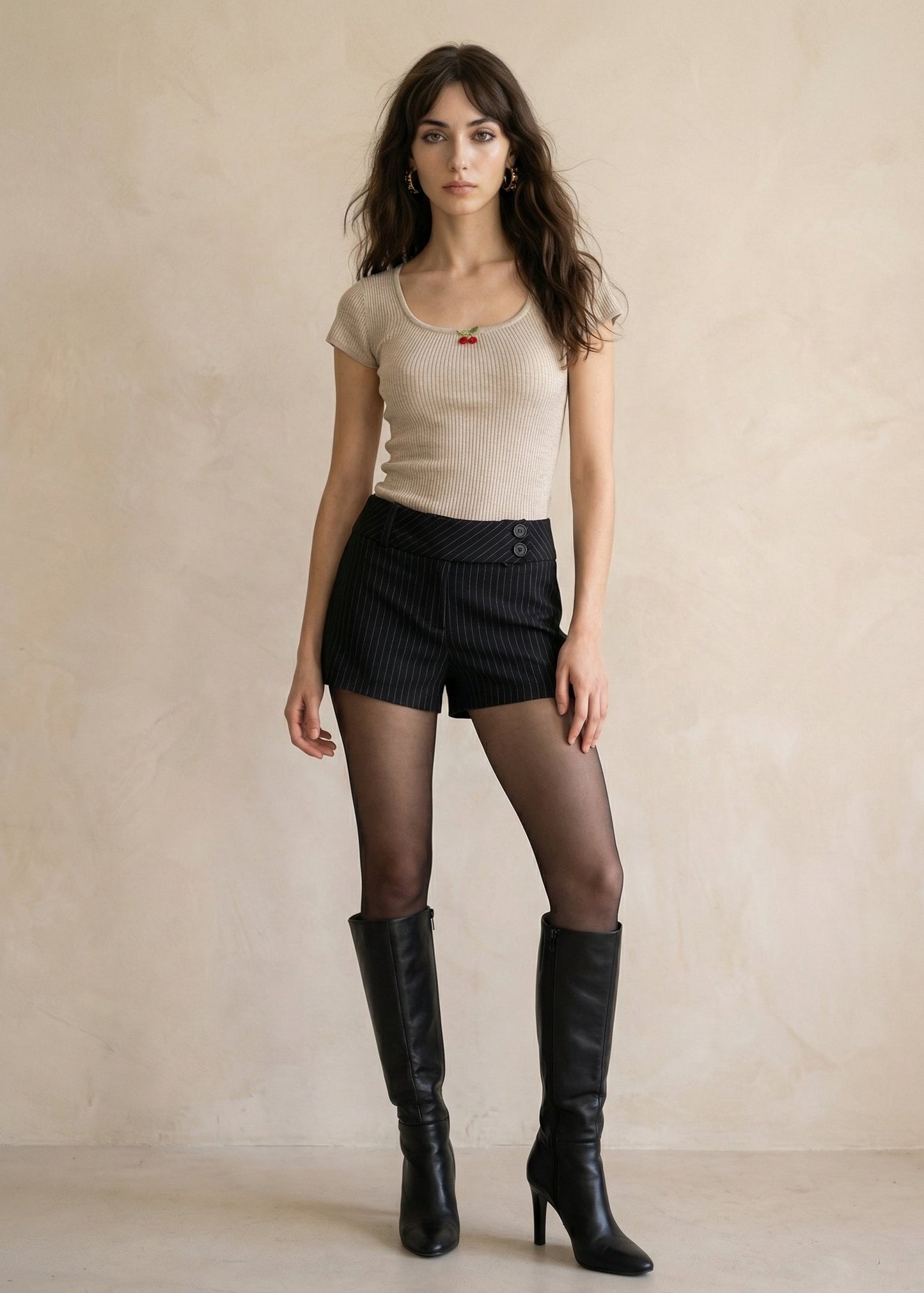 Woman wearing a beige top, black shorts, and black knee-high boots against a plain background