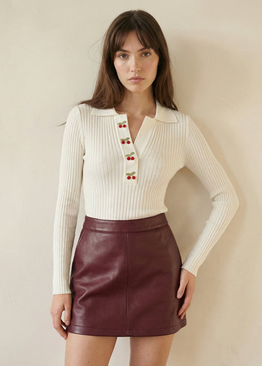 Woman wearing a cream ribbed sweater with red button details and a maroon leather skirt against a beige background