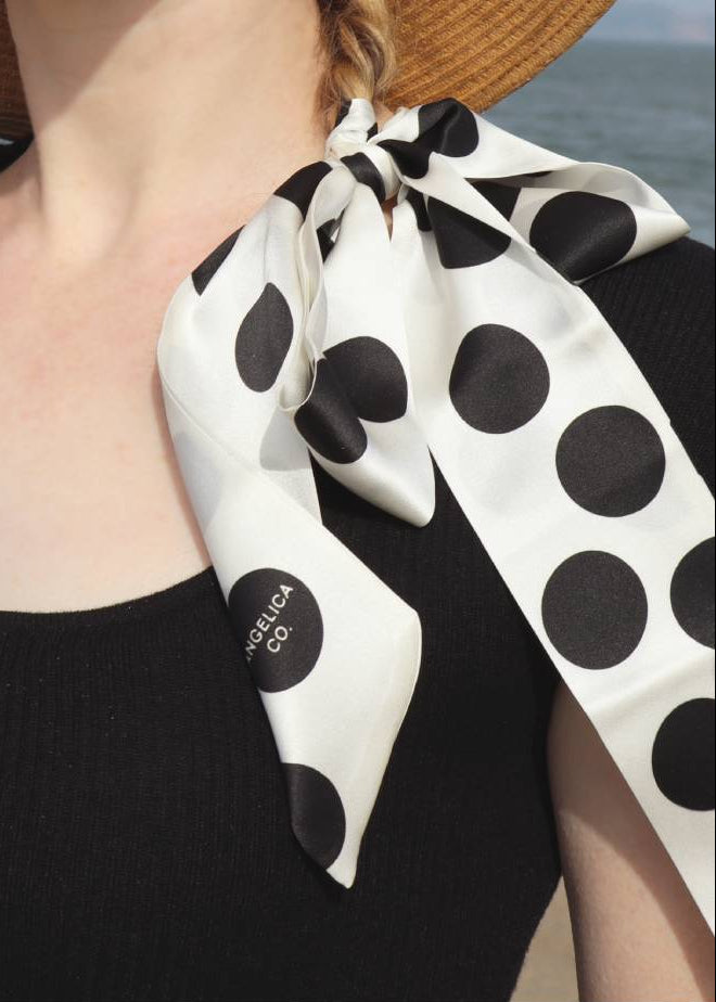 Person wearing a black tank top with cherry design and a polka dot scarf, standing by water.