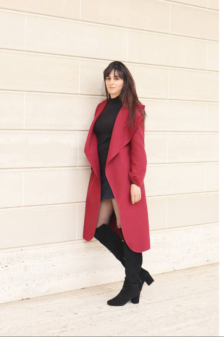 Person wearing a red coat and black outfit standing against a light-colored wall.