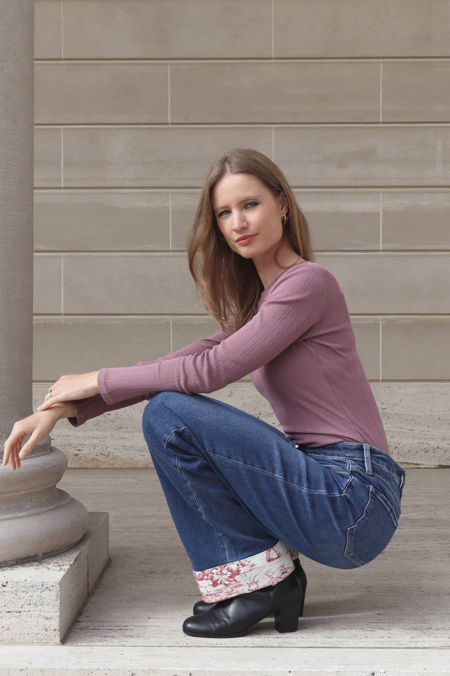 Woman in a pink top and blue jeans sitting on stone steps against a beige wall.