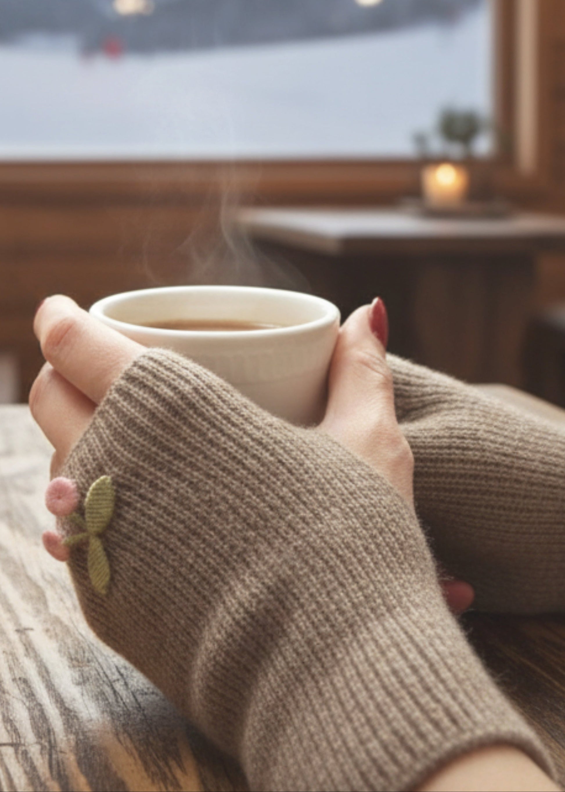 Person holding a steaming cup of coffee with knitted sleeves in front of a window with string lights and mountains.