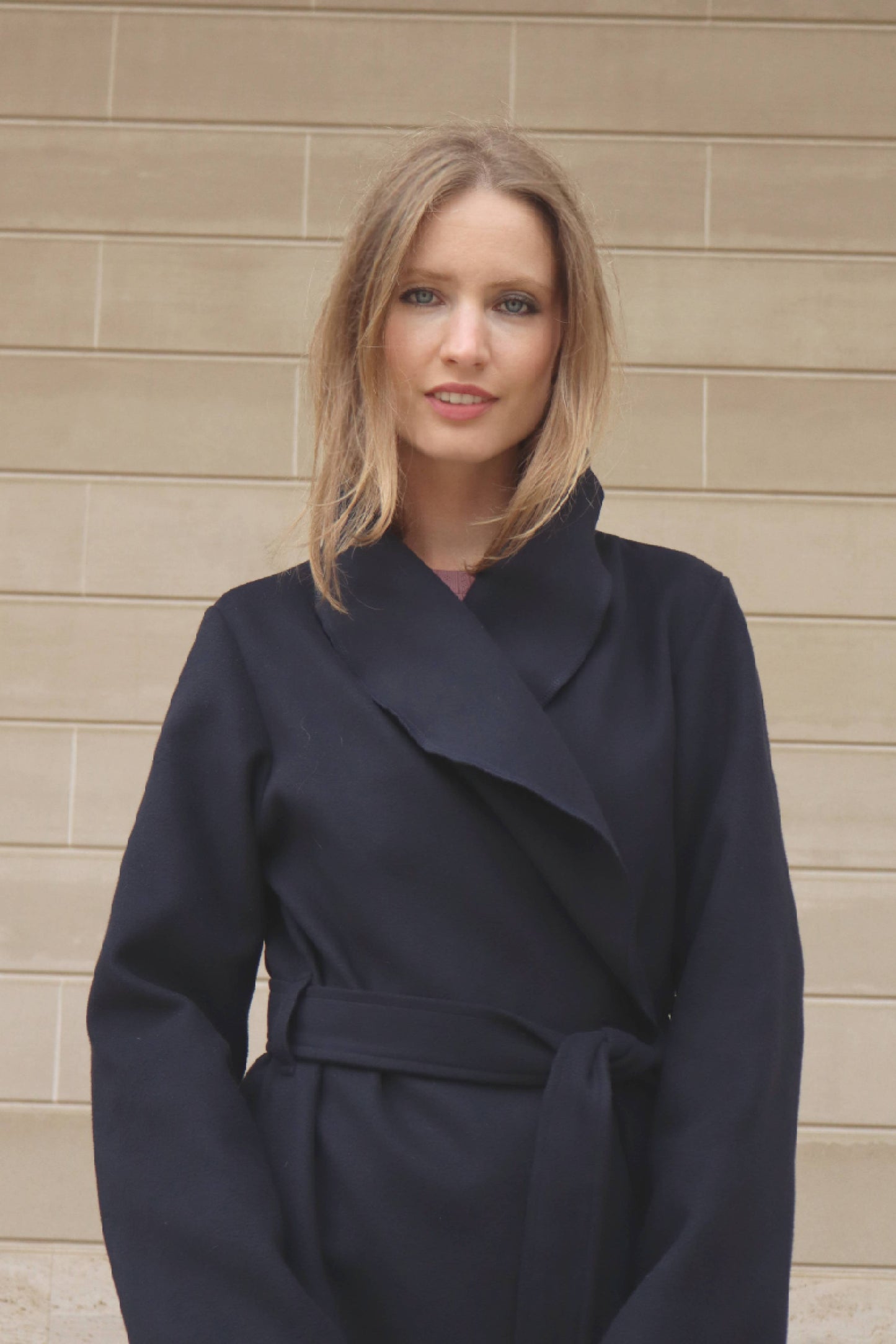 Woman wearing a navy blue coat standing against a beige wall.