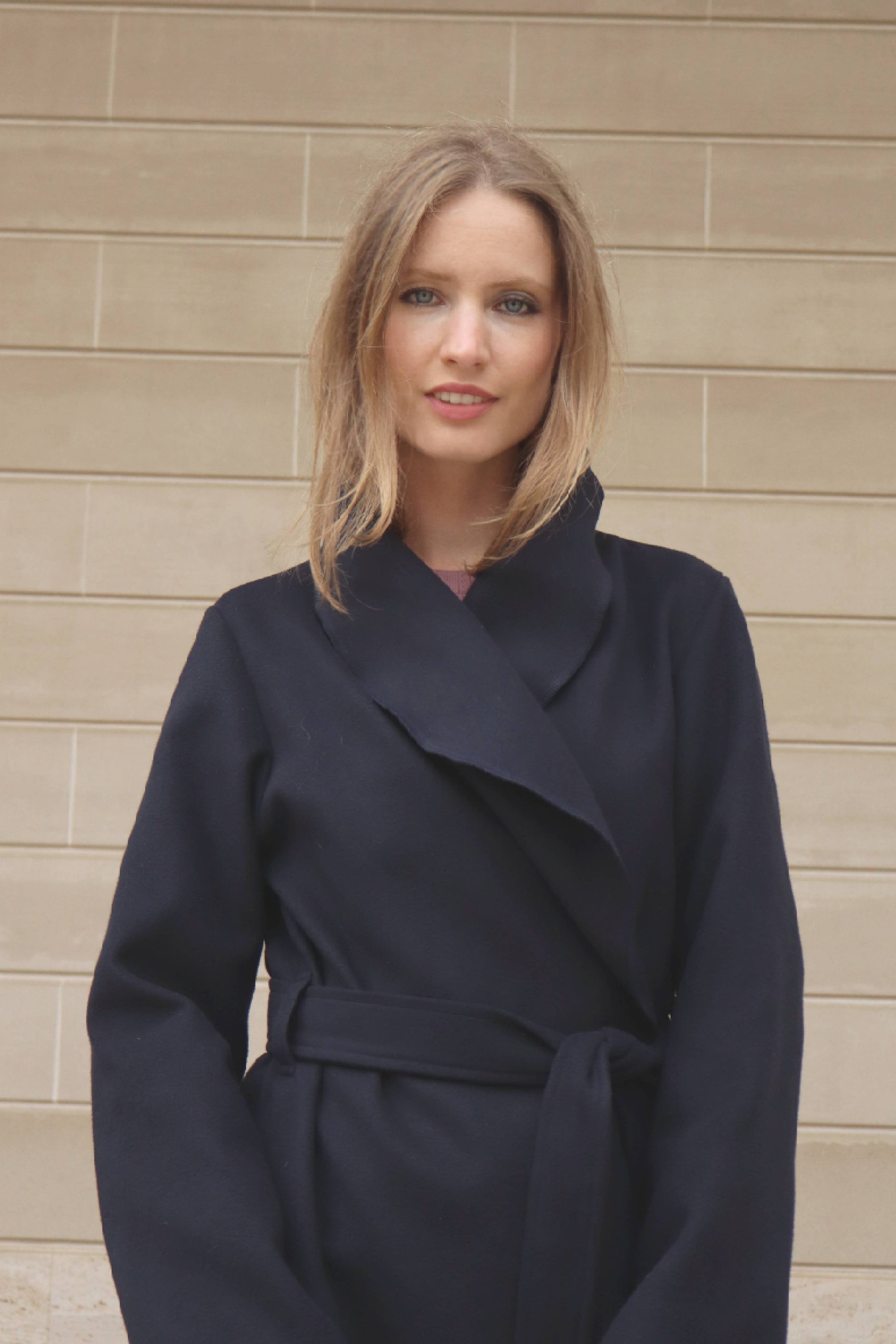 Woman wearing a navy blue coat standing against a beige wall.