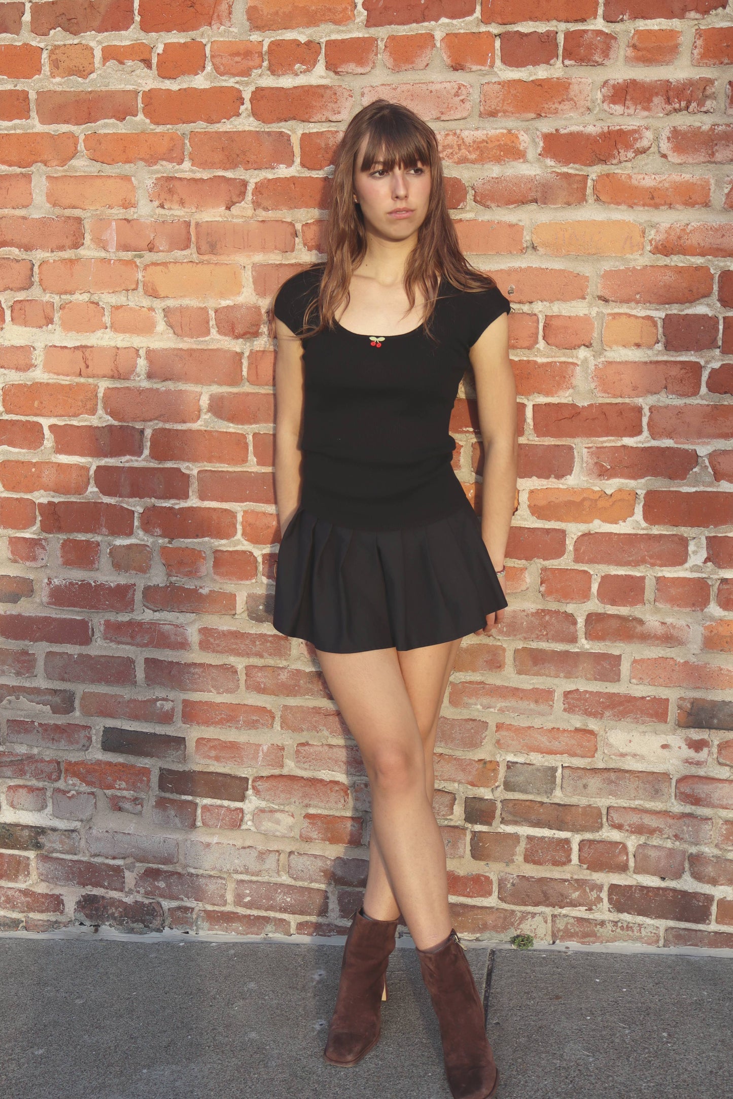 Woman in a black dress and brown boots standing against a brick wall.