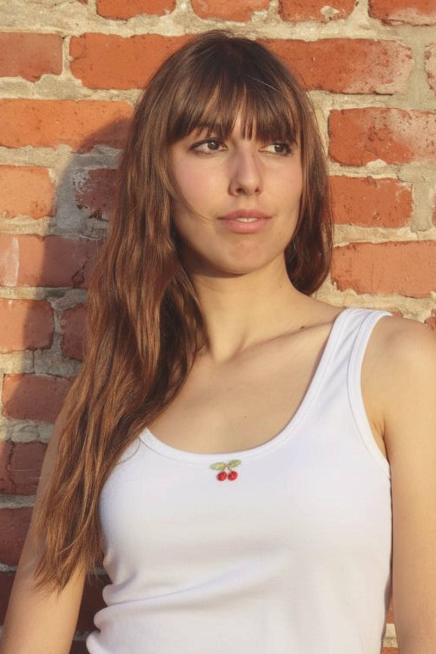 Woman wearing a white tank top with cherry design against a brick wall